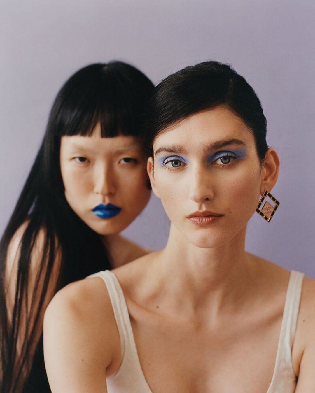 Two women pose in a close-up shot, showcasing bold blue lipstick and eyeshadow against a light purple backdrop. Beauty and fashion portrait.