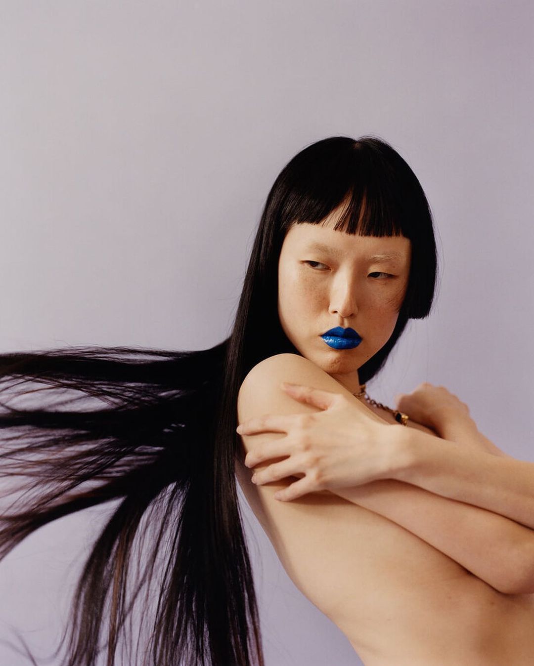 Asian woman with black hair and blue lipstick poses topless against a light purple backdrop, highlighting makeup and hairstyle in a studio shot.