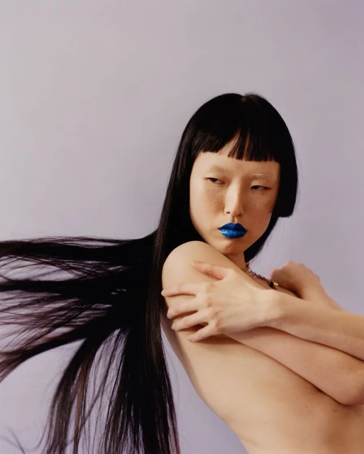 Asian woman with black hair and blue lipstick poses topless against a light purple backdrop, highlighting makeup and hairstyle in a studio shot.
