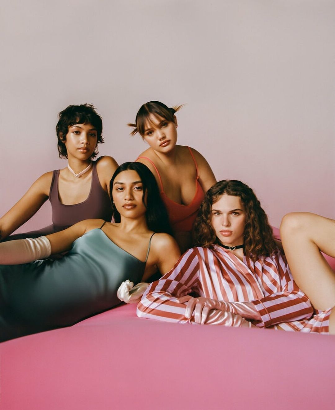 Four female fashion models in vintage-inspired attire pose on a pink backdrop in a studio setting with soft, diffused lighting.