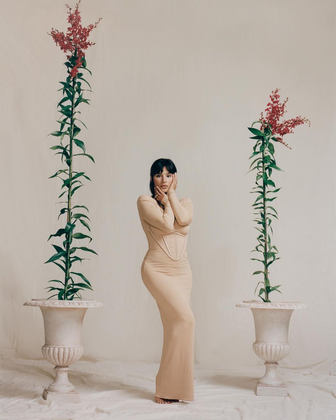 Woman in beige dress poses between two floral arrangements in urns against a light beige background in a fashion shot.