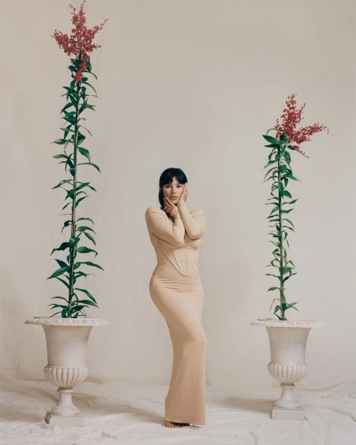 Woman in beige dress poses between two floral arrangements in urns against a light beige background in a fashion shot.