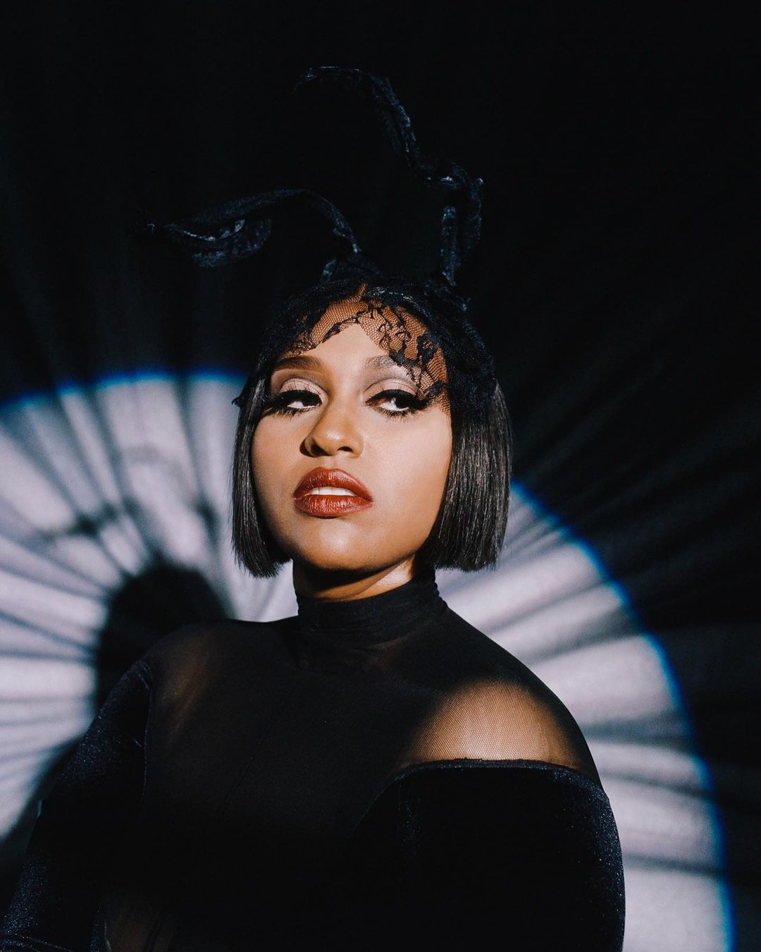Portrait of a woman with dark hair, wearing a black turtleneck and headpiece, looking up against a dark background with lighting effects.
