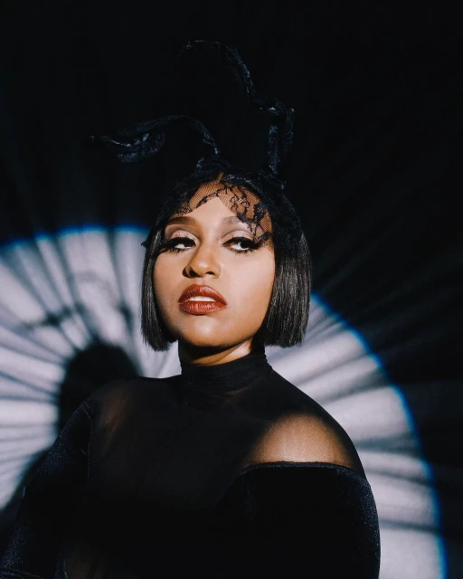 Portrait of a woman with dark hair, wearing a black turtleneck and headpiece, looking up against a dark background with lighting effects.
