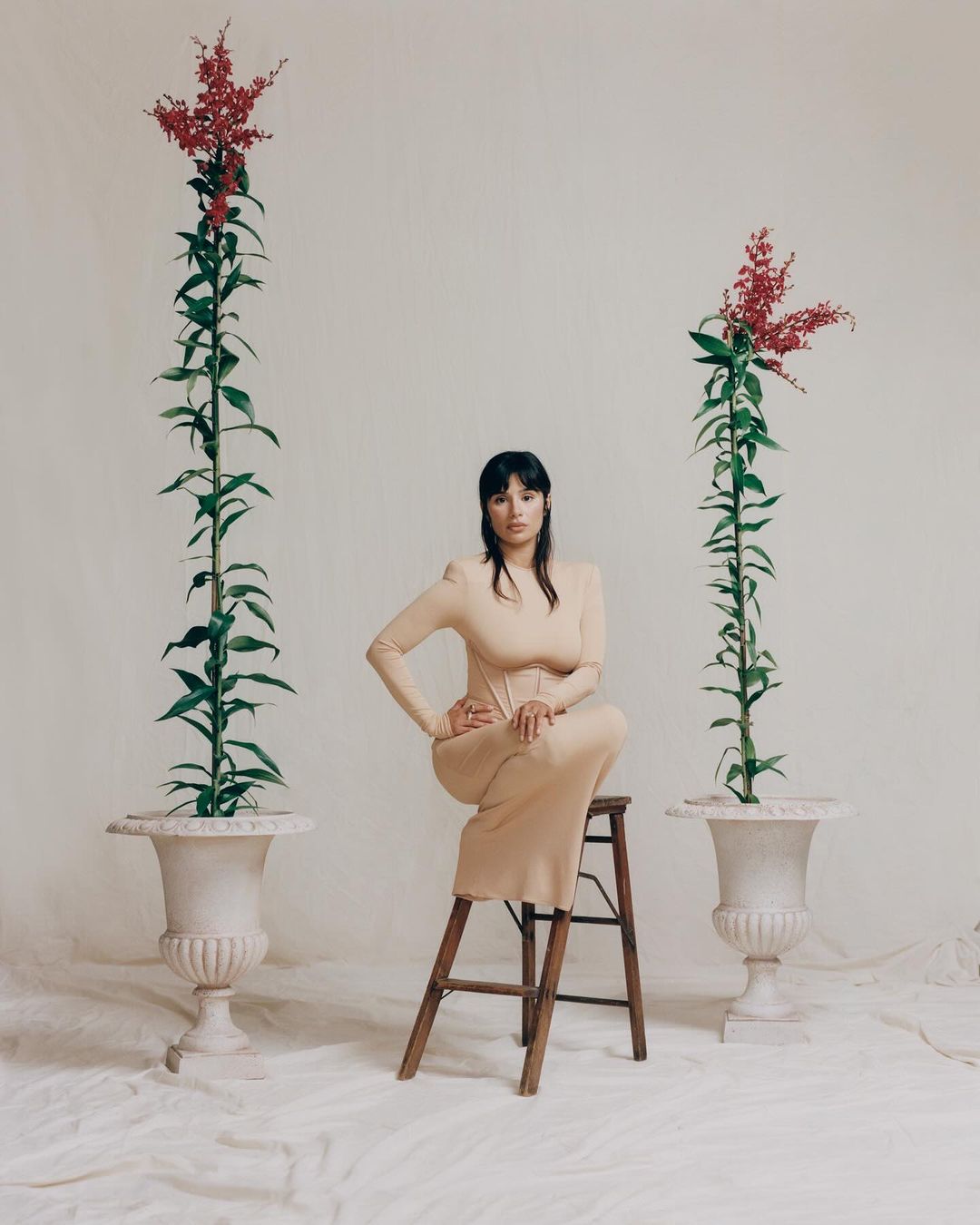 Woman in beige dress sits on wooden stool between two plants in urns, set against off-white background in a fashion editorial.
