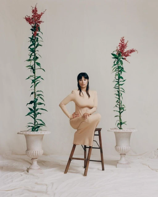 Woman in beige dress sits on wooden stool between two plants in urns, set against off-white background in a fashion editorial.