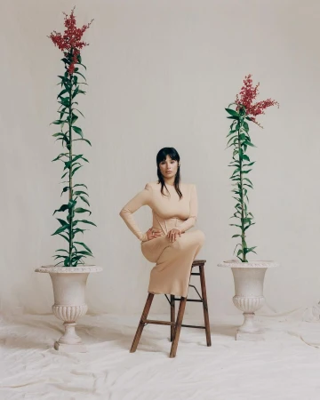 Woman in beige dress sits on wooden stool between two plants in urns, set against off-white background in a fashion editorial.