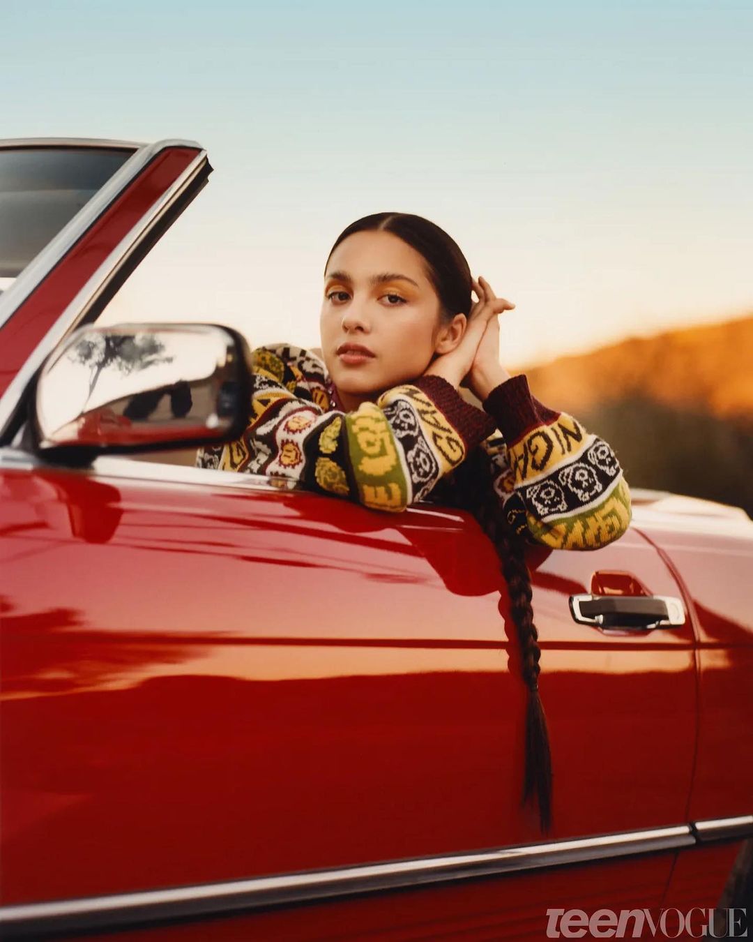 Portrait of a young woman in a red convertible car, wearing patterned sweater with braided hair, set against a clear blue sky.