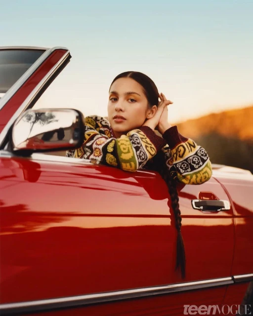 Portrait of a young woman in a red convertible car, wearing patterned sweater with braided hair, set against a clear blue sky.