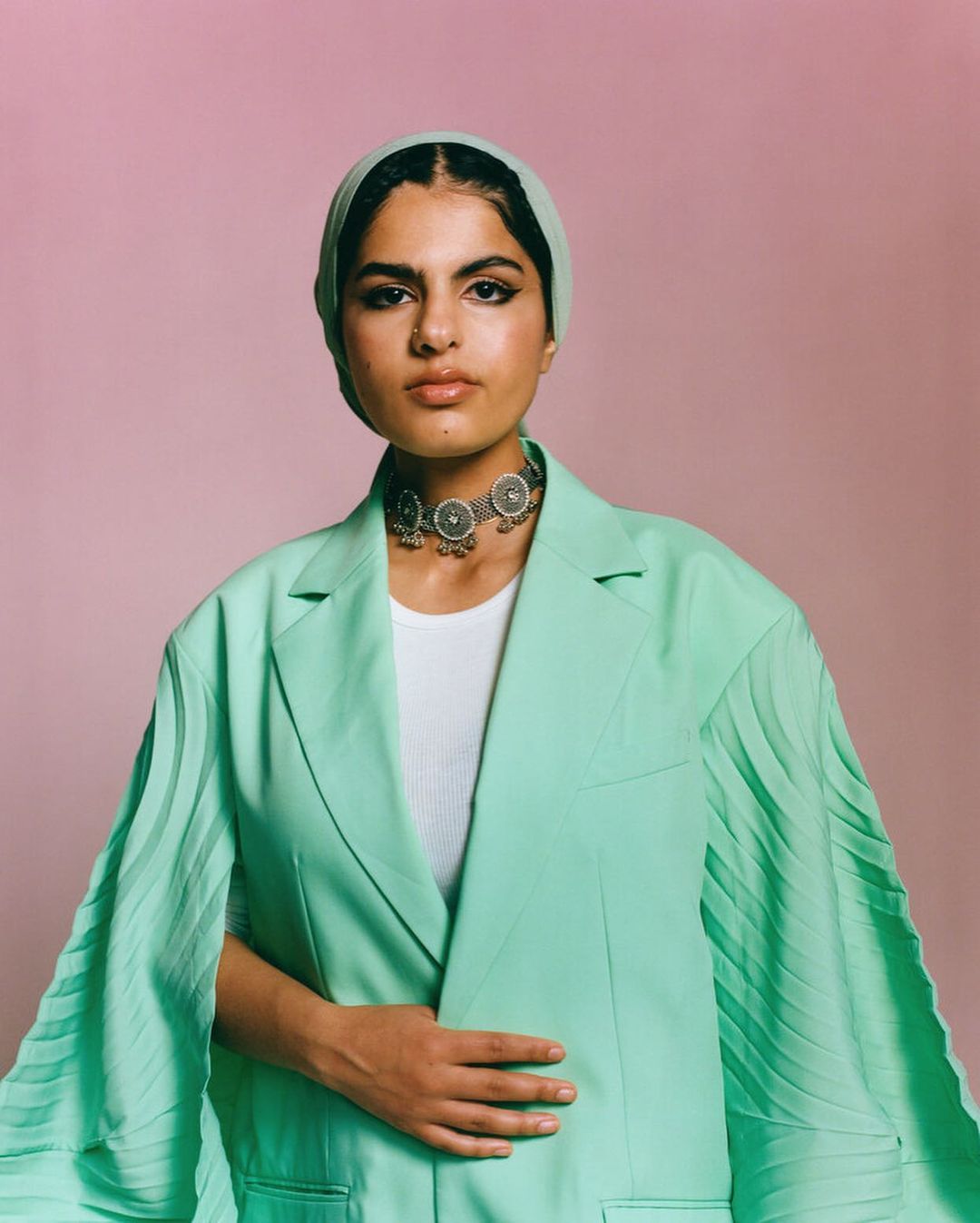Woman in headscarf and mint green blazer poses against a pink background, showcasing a unique silver necklace and confident expression.