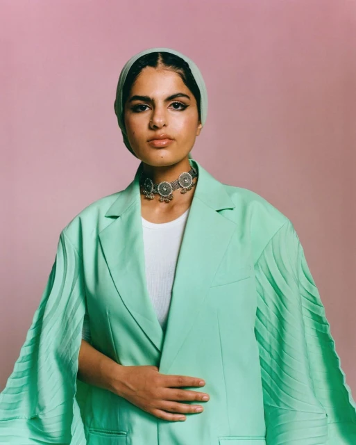 Woman in headscarf and mint green blazer poses against a pink background, showcasing a unique silver necklace and confident expression.