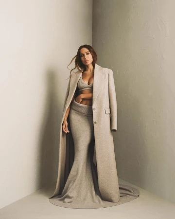 Elegant adult woman in a knitted bra top and long skirt, paired with a long coat in a minimalist indoor setting with neutral tones.