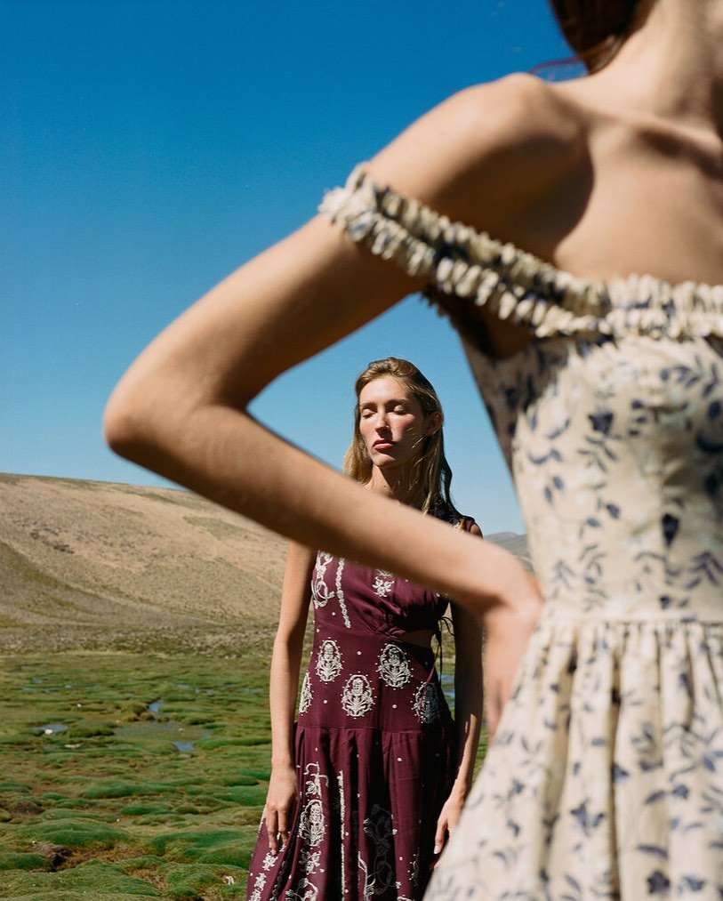 Two women modeling patterned dresses in an open landscape with sparse green vegetation and a bright blue sky.