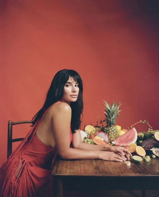 Attractive brunette woman poses at a wooden table laden with fresh fruit against a solid red background in a studio portrait.