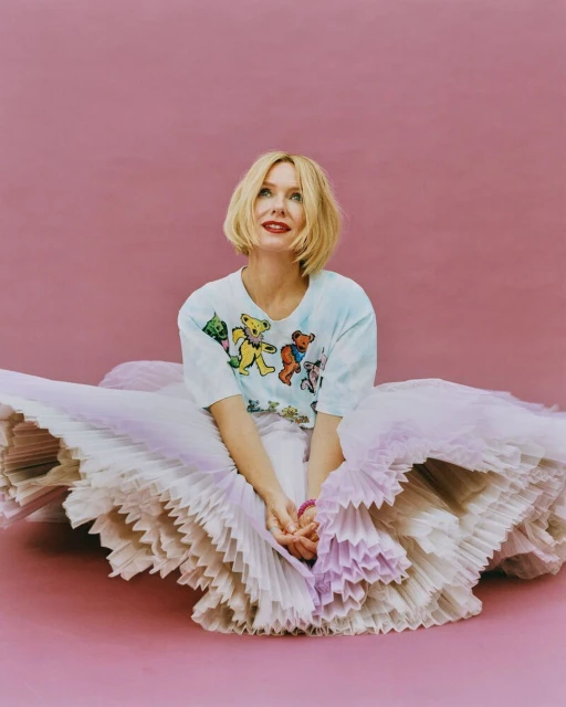 Woman in graphic t-shirt and layered tulle skirt poses against pink backdrop in eye-level studio portrait. Fashion and entertainment.