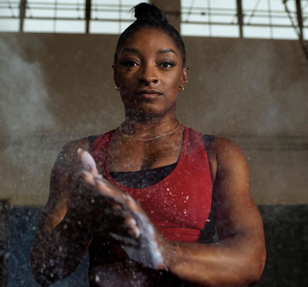 Portrait of Simone Biles with chalk on hands, facing forward in workout gear, shot in eye-level medium shot, in neutral indoor setting.