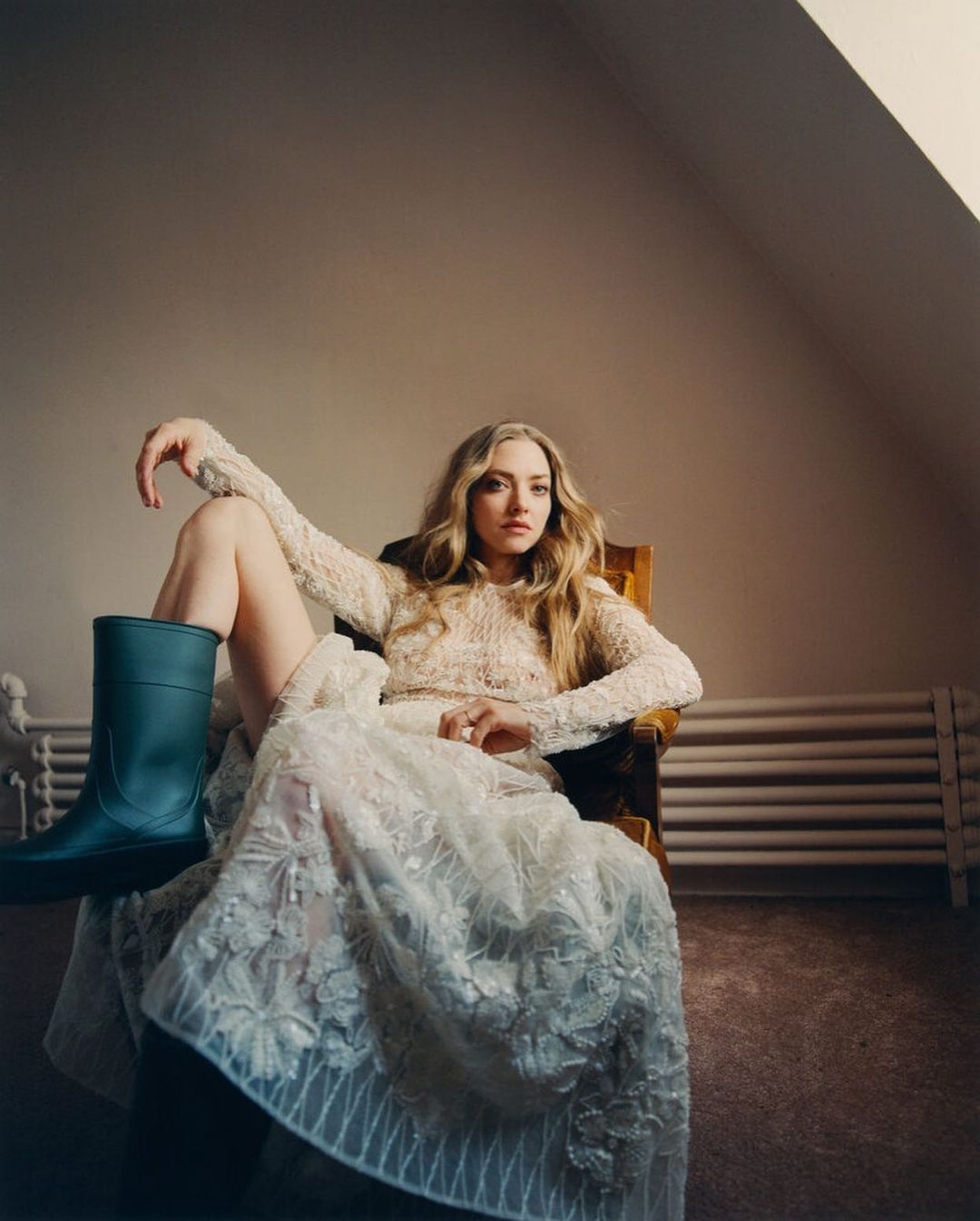 Amanda Seyfried poses in a chair wearing a floral dress and teal rain boot in an indoor setting with neutral color palette.