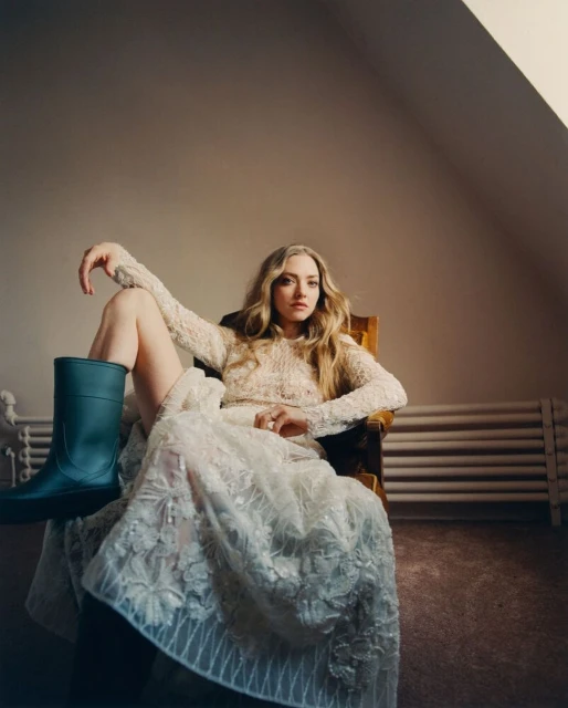 Amanda Seyfried poses in a chair wearing a floral dress and teal rain boot in an indoor setting with neutral color palette.