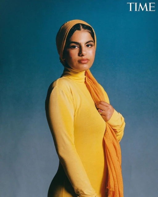 Portrait of a woman wearing a yellow turtleneck and headscarf, posing against a blue background. The 'TIME' logo is visible in the corner.