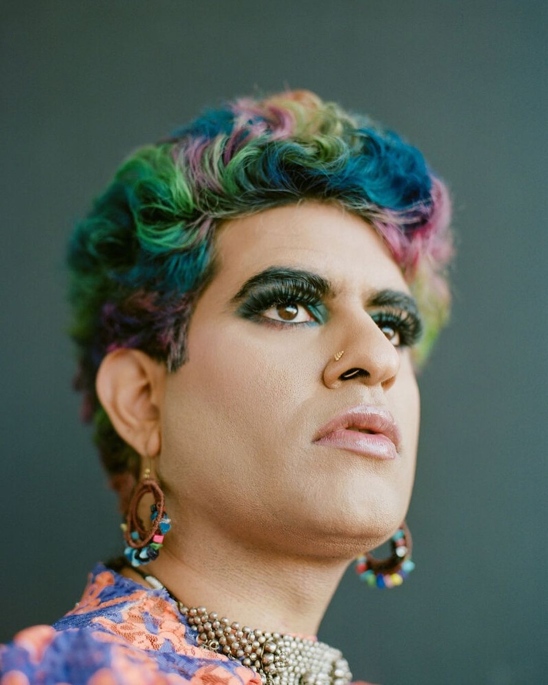 Portrait of a person with rainbow-colored hair, heavy eye makeup, a nose ring, earrings, and a beaded necklace, looking up in a studio.