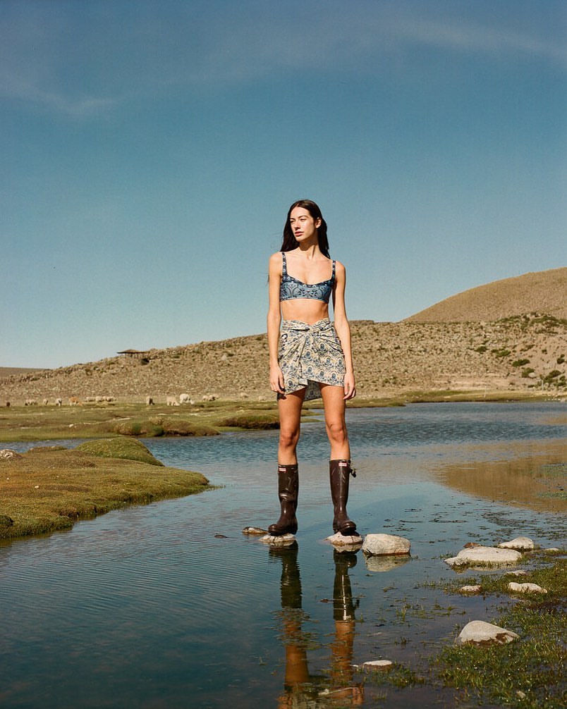 A woman in a bikini and sarong stands on rocks in a body of water in a rural landscape on a clear, sunny day, fashion photography.