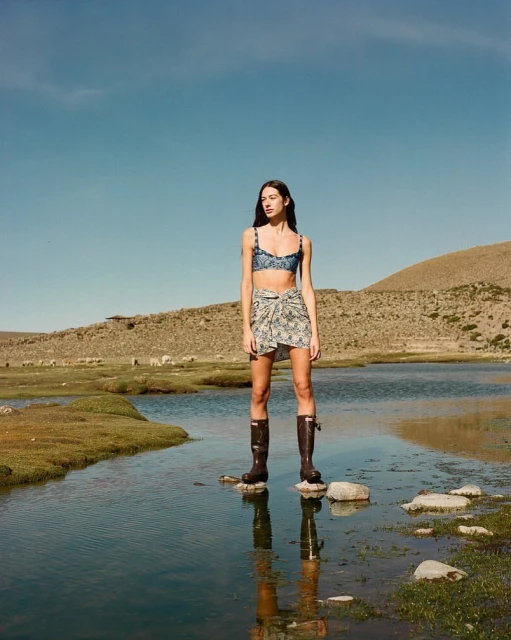 A woman in a bikini and sarong stands on rocks in a body of water in a rural landscape on a clear, sunny day, fashion photography.