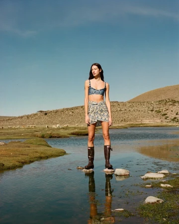 A woman in a bikini and sarong stands on rocks in a body of water in a rural landscape on a clear, sunny day, fashion photography.