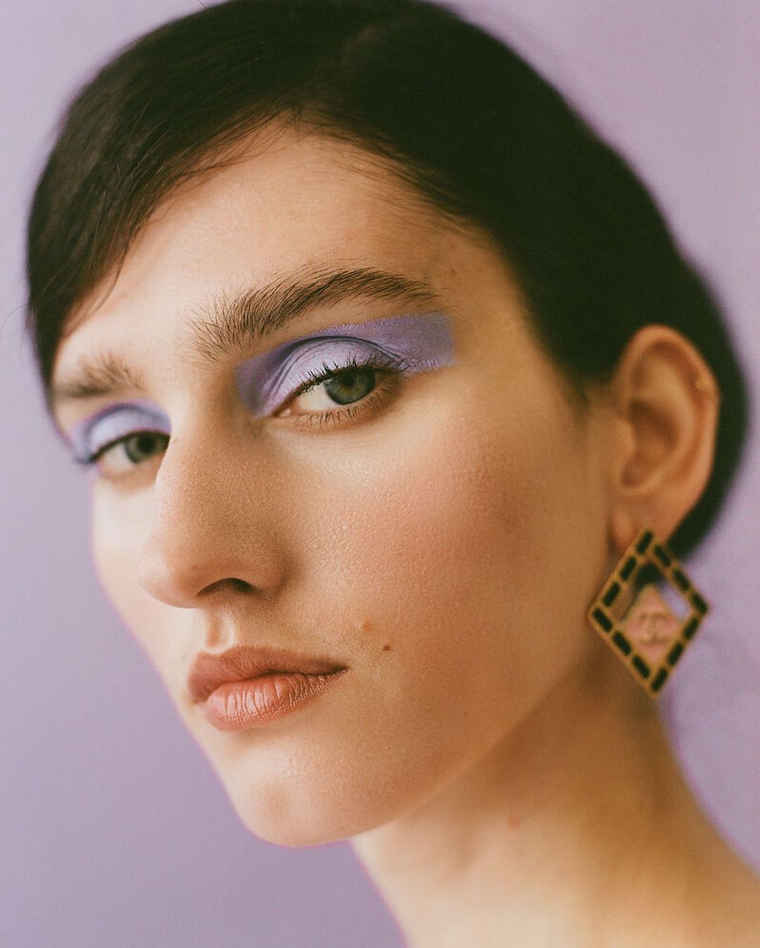 Close-up of a woman with purple eye shadow and large square earrings against a lilac background in a beauty-style portrait.