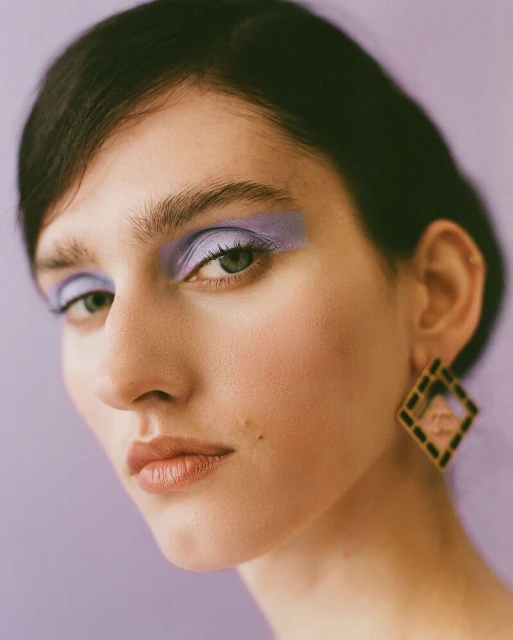 Close-up of a woman with purple eye shadow and large square earrings against a lilac background in a beauty-style portrait.