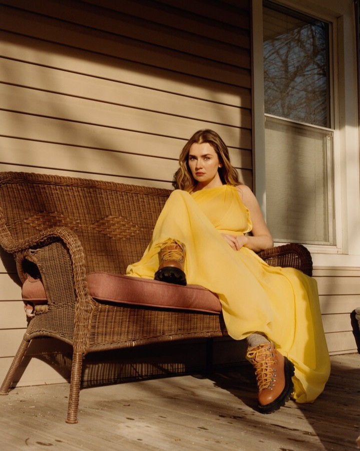 Woman in a yellow dress and hiking boots sits on a wicker bench outside a house with wood siding in the late afternoon sunlight.