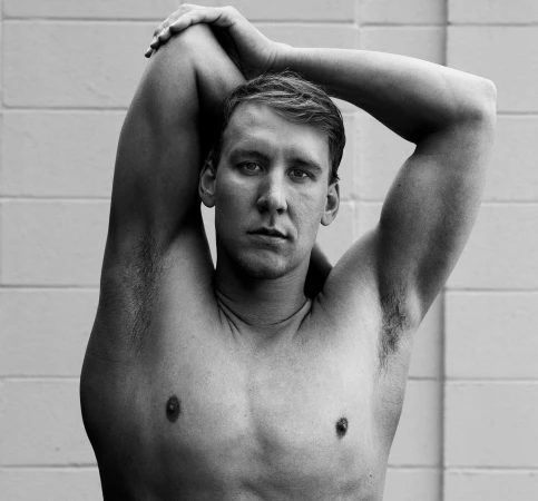 Black and white portrait of a shirtless man with arms raised and hands clasped behind his head against a brick wall.