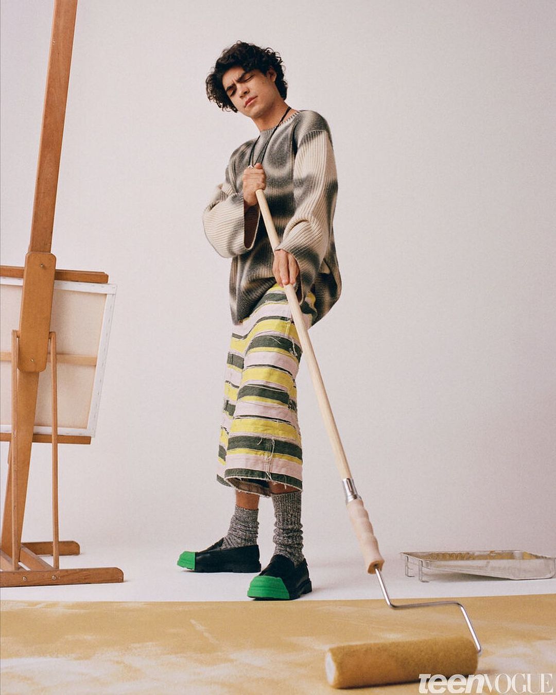 Young adult man in patterned sweater and shorts holds paint roller in studio, posing near canvas and paint tray; studio lighting.