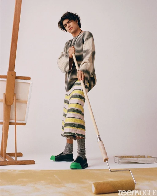 Young adult man in patterned sweater and shorts holds paint roller in studio, posing near canvas and paint tray; studio lighting.