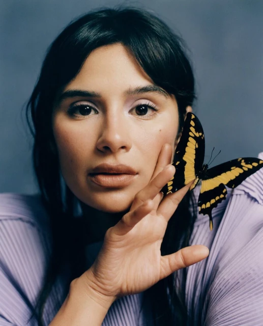 Close-up portrait of a woman with dark hair and light skin, wearing a lilac top, with a large black and yellow butterfly on her face.