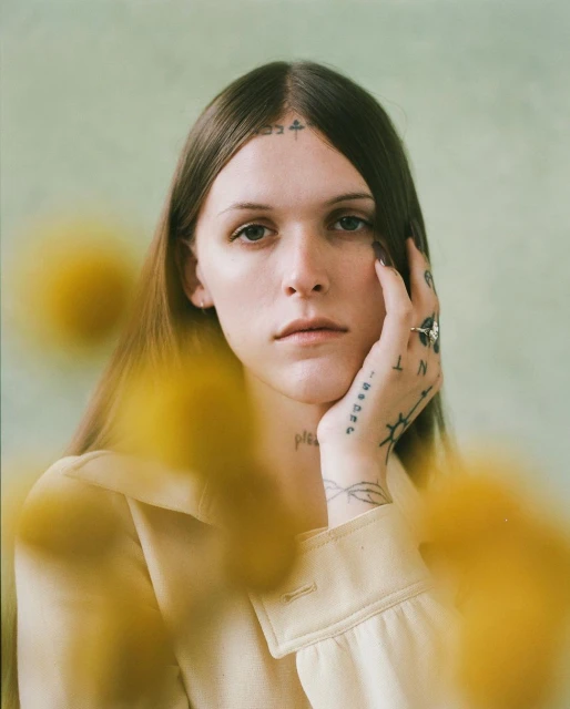 Portrait of a person with long brown hair, wearing a yellow collared shirt with visible tattoos and a ring, posing in front of a blurred green background.