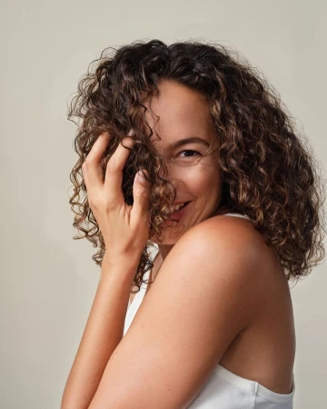 Portrait of a smiling woman with dark curly hair wearing a white top against a neutral background, photographed by Josefina Santos.