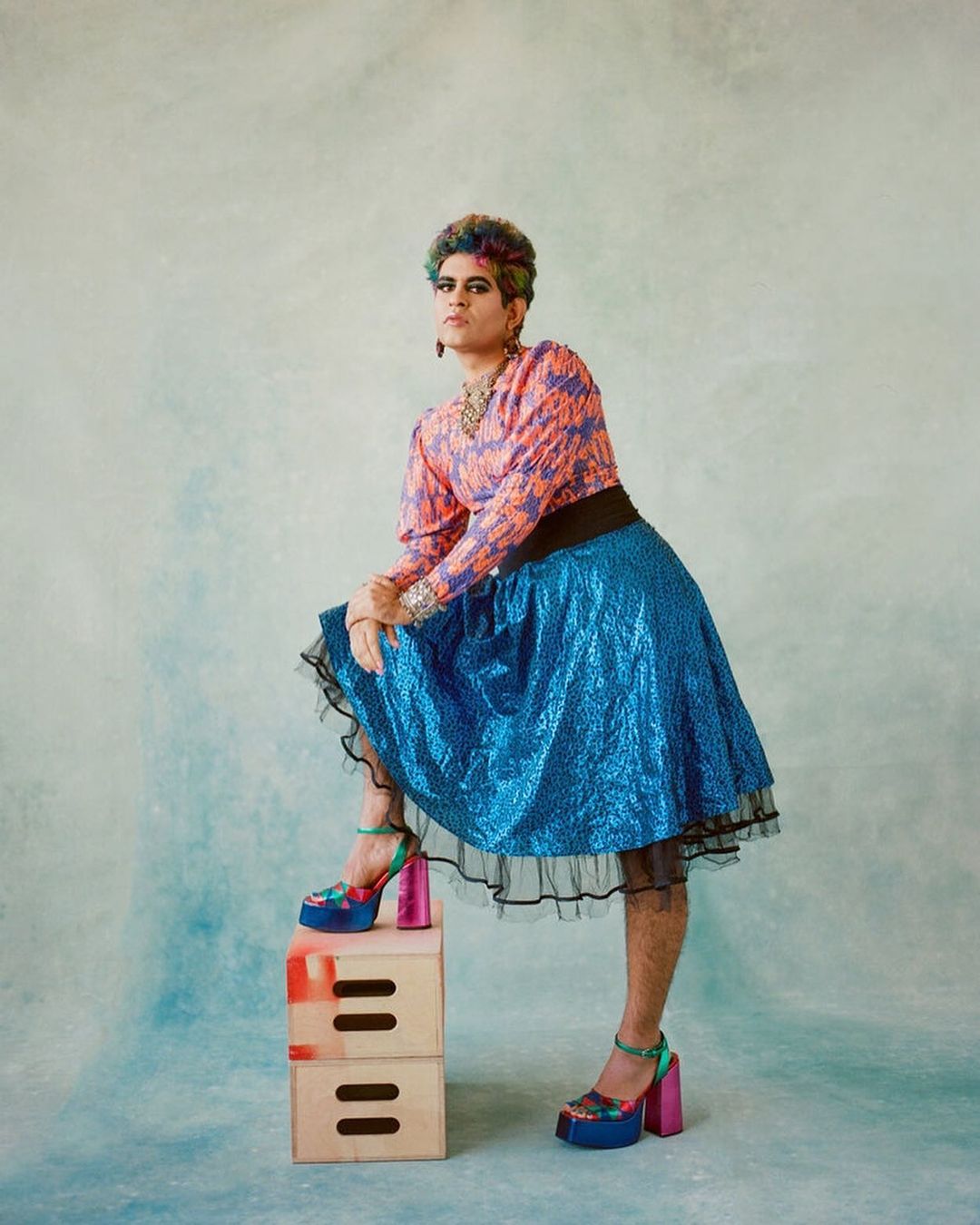 Person in pink top and blue skirt posing on wooden crates against a mottled backdrop, accessorized with jewelry and platform heels.