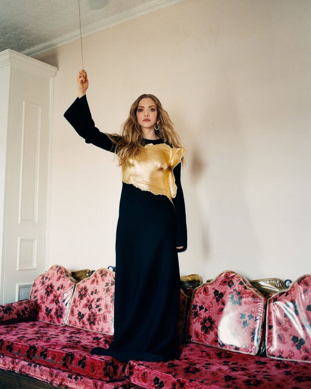 Amanda Seyfried poses indoors on a floral sofa, wearing a long black dress with a gold top, in a full-length portrait.