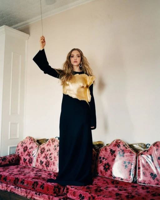Amanda Seyfried poses indoors on a floral sofa, wearing a long black dress with a gold top, in a full-length portrait.