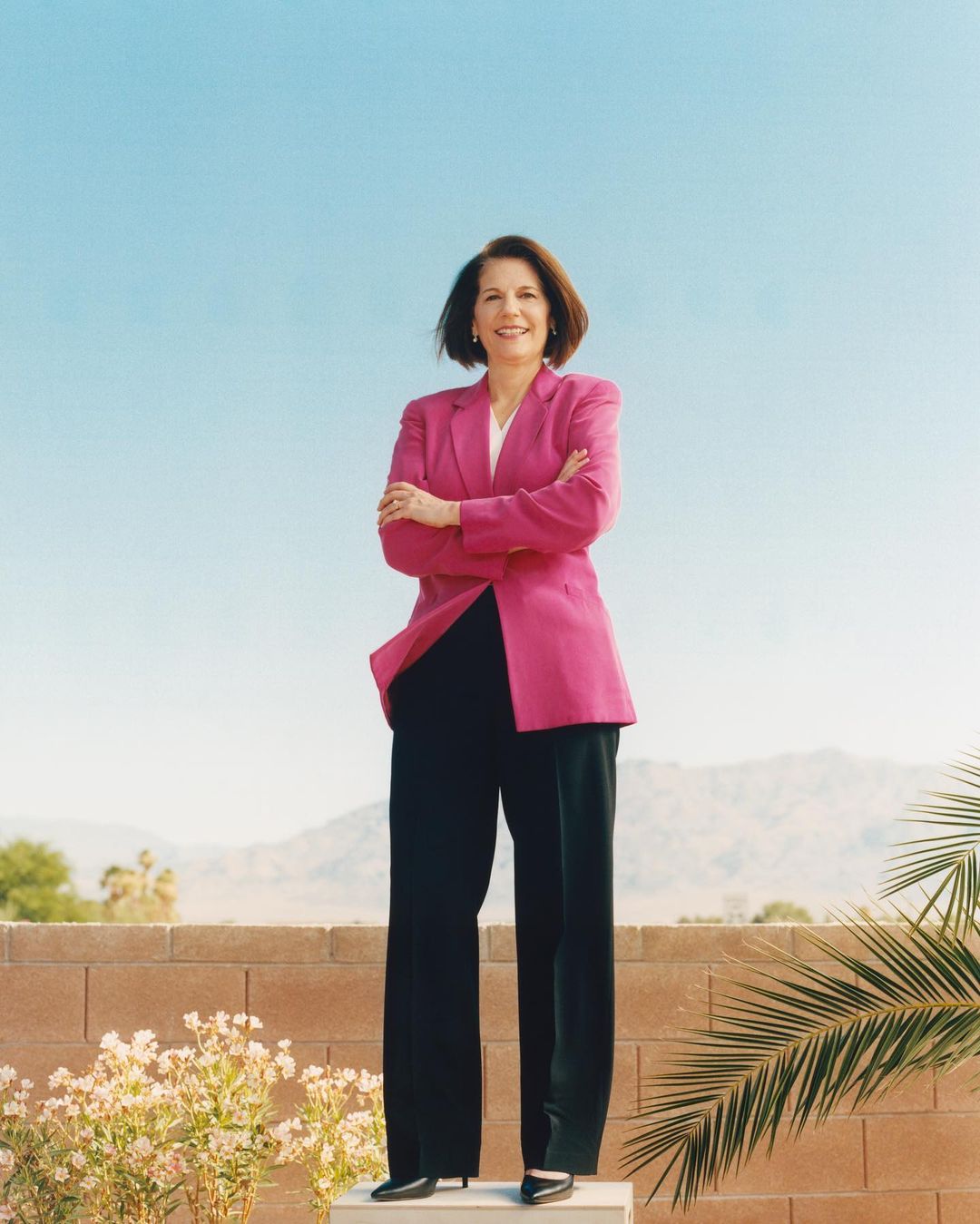 Full shot of a smiling, middle-aged woman in a bright pink blazer and black pants standing outdoors with mountains in the background.