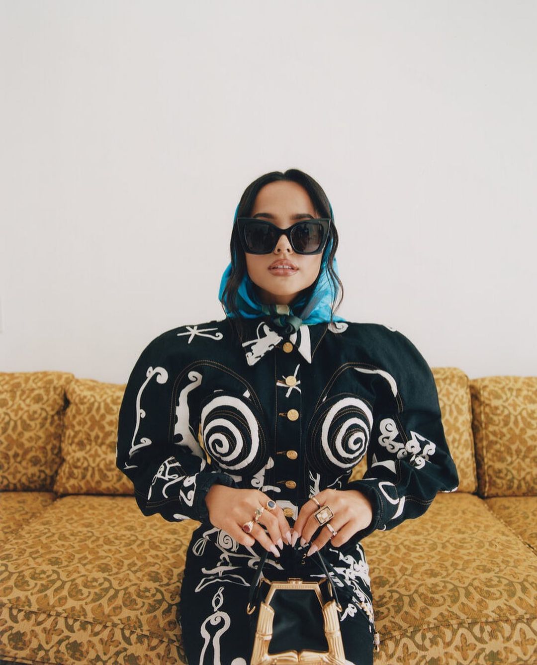 A woman with sunglasses and a blue headscarf poses indoors wearing a black and white patterned jacket and dress with a yellow couch behind her.
