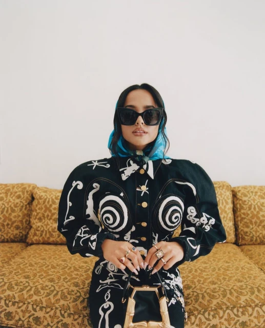 A woman with sunglasses and a blue headscarf poses indoors wearing a black and white patterned jacket and dress with a yellow couch behind her.