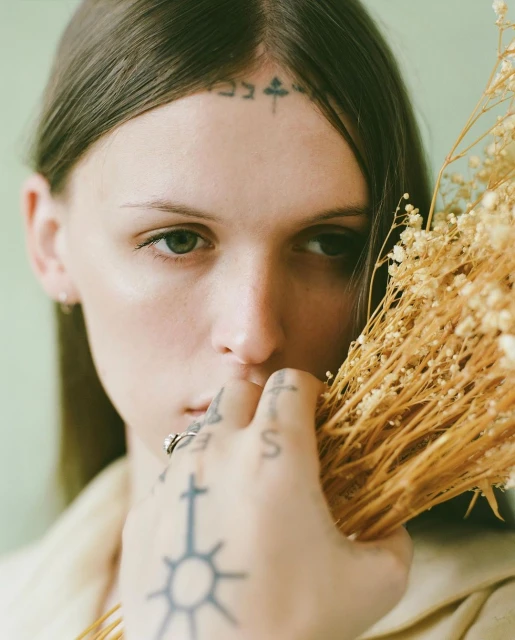 Close-up portrait of a young woman with tattoos, holding dried flowers, wearing a cream-colored jacket against a pale green background.