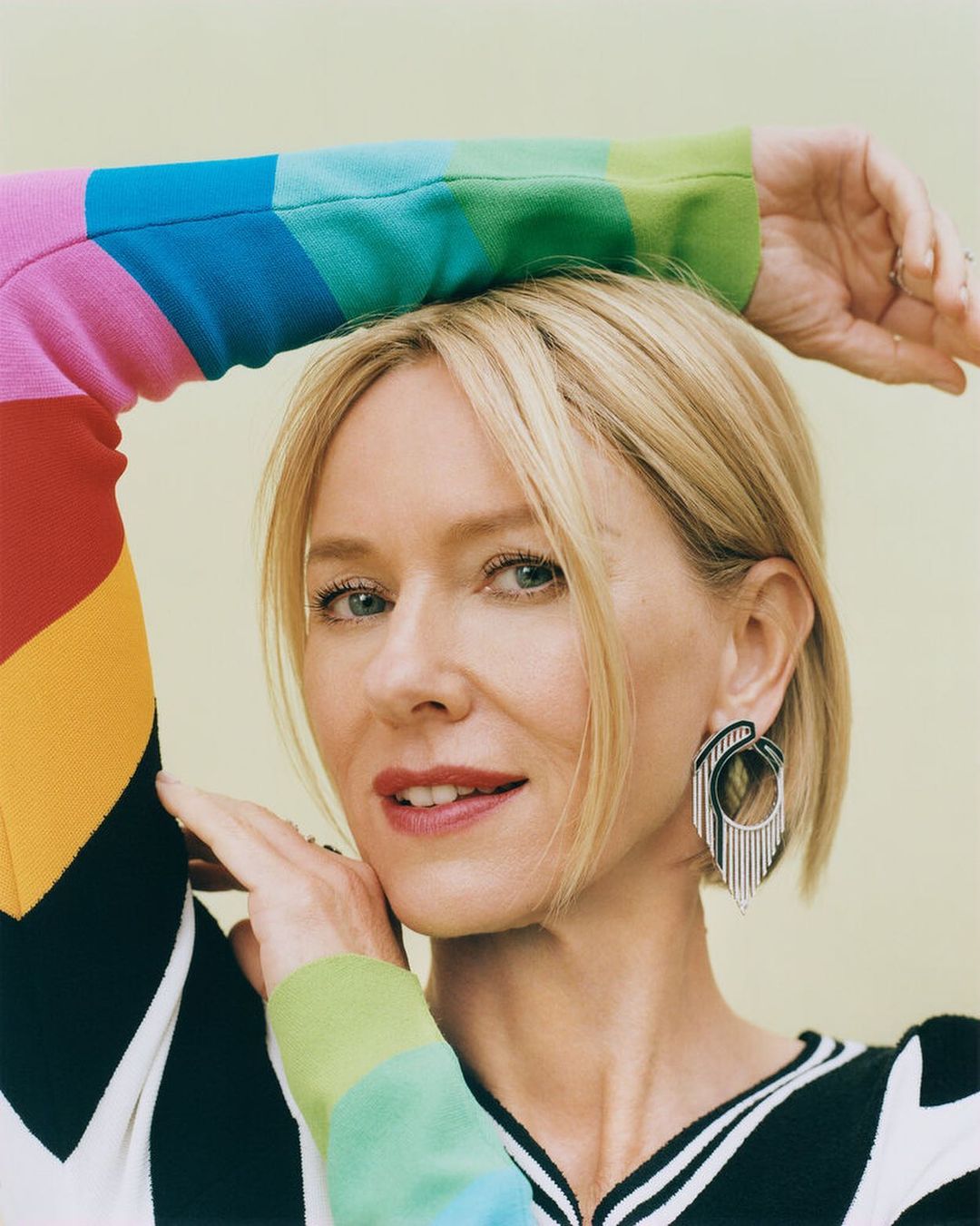Portrait of a blonde woman with an arm raised, wearing a rainbow-colored sweater and statement earrings against a light yellow backdrop.