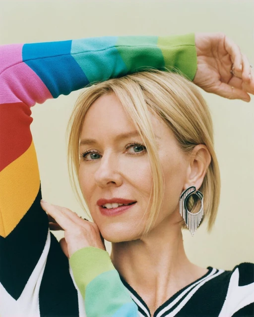 Portrait of a blonde woman with an arm raised, wearing a rainbow-colored sweater and statement earrings against a light yellow backdrop.