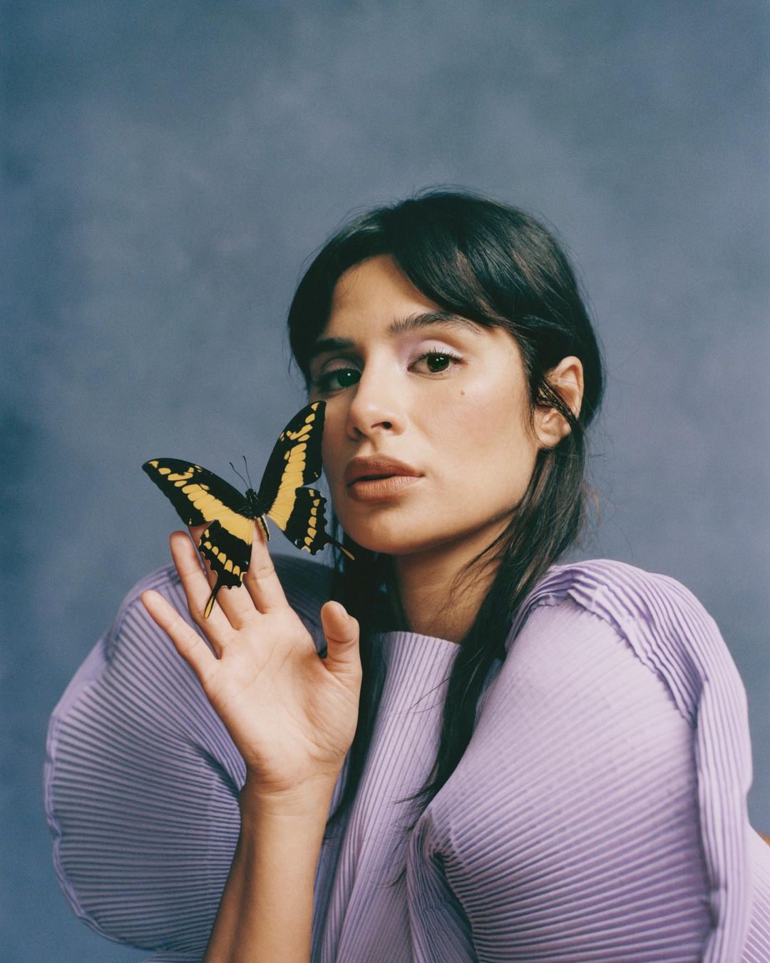 Portrait of a woman with dark hair and a purple top, holding a yellow and black butterfly on her hand against a mottled blue background.