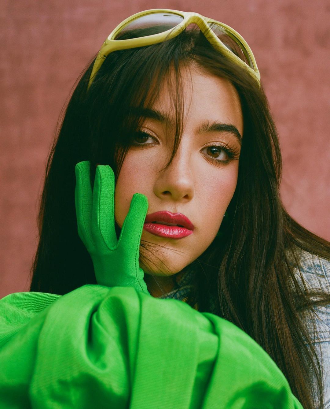 Close-up portrait of a woman wearing bright green gloves, a vivid green top, and yellow sunglasses resting on her dark hair.