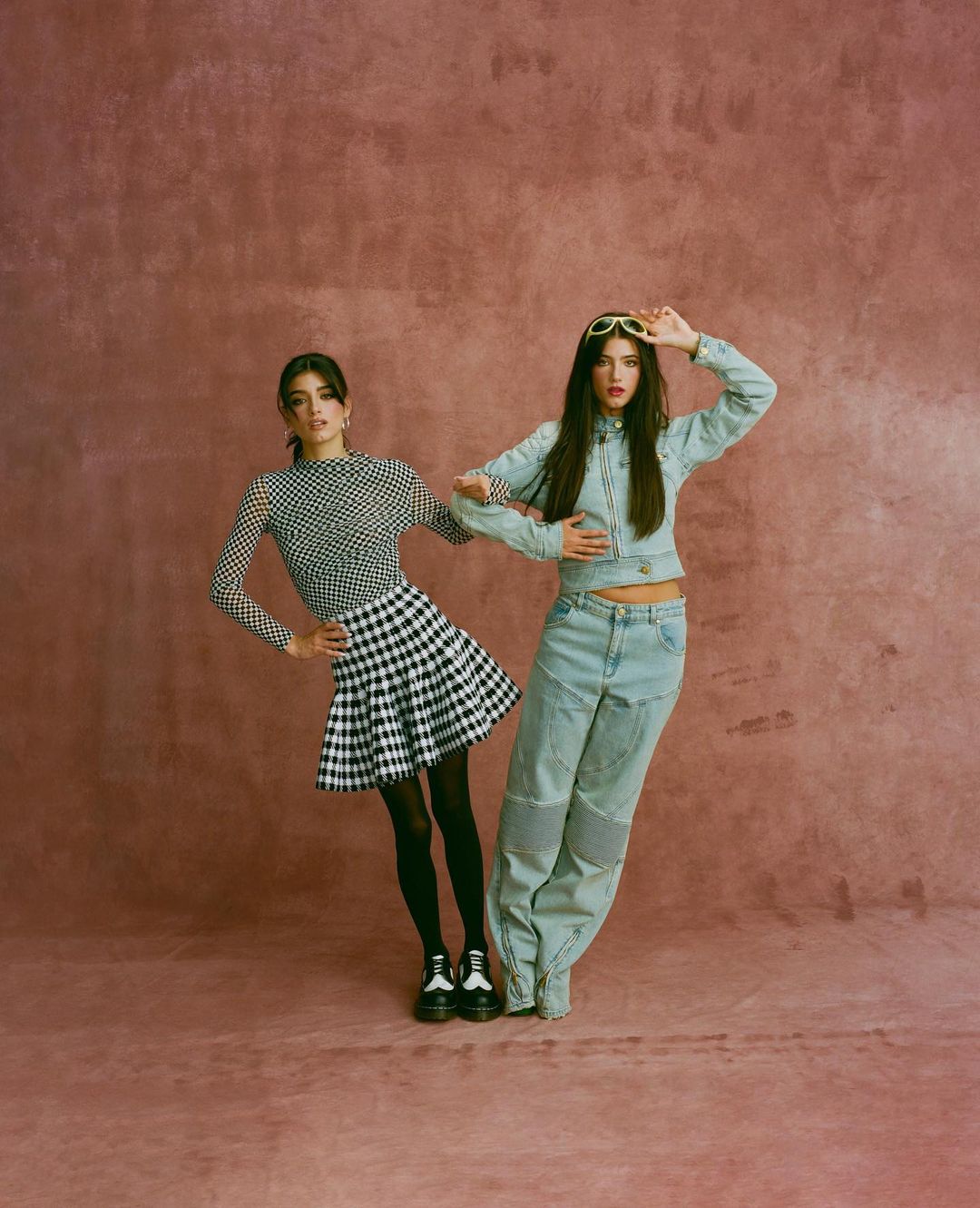 Two women pose in a photo studio, one in a gingham skirt and the other in a denim outfit, against a textured pink backdrop.