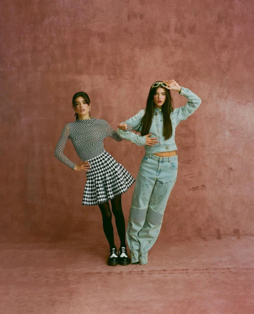Two women pose in a photo studio, one in a gingham skirt and the other in a denim outfit, against a textured pink backdrop.