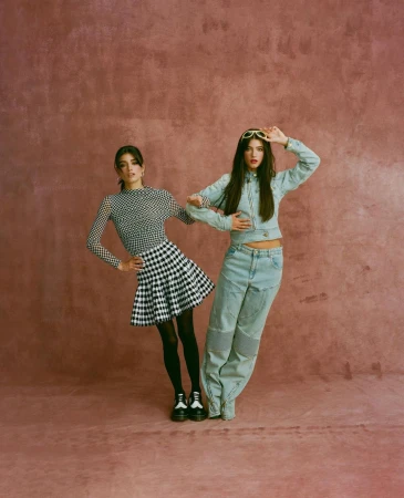 Two women pose in a photo studio, one in a gingham skirt and the other in a denim outfit, against a textured pink backdrop.